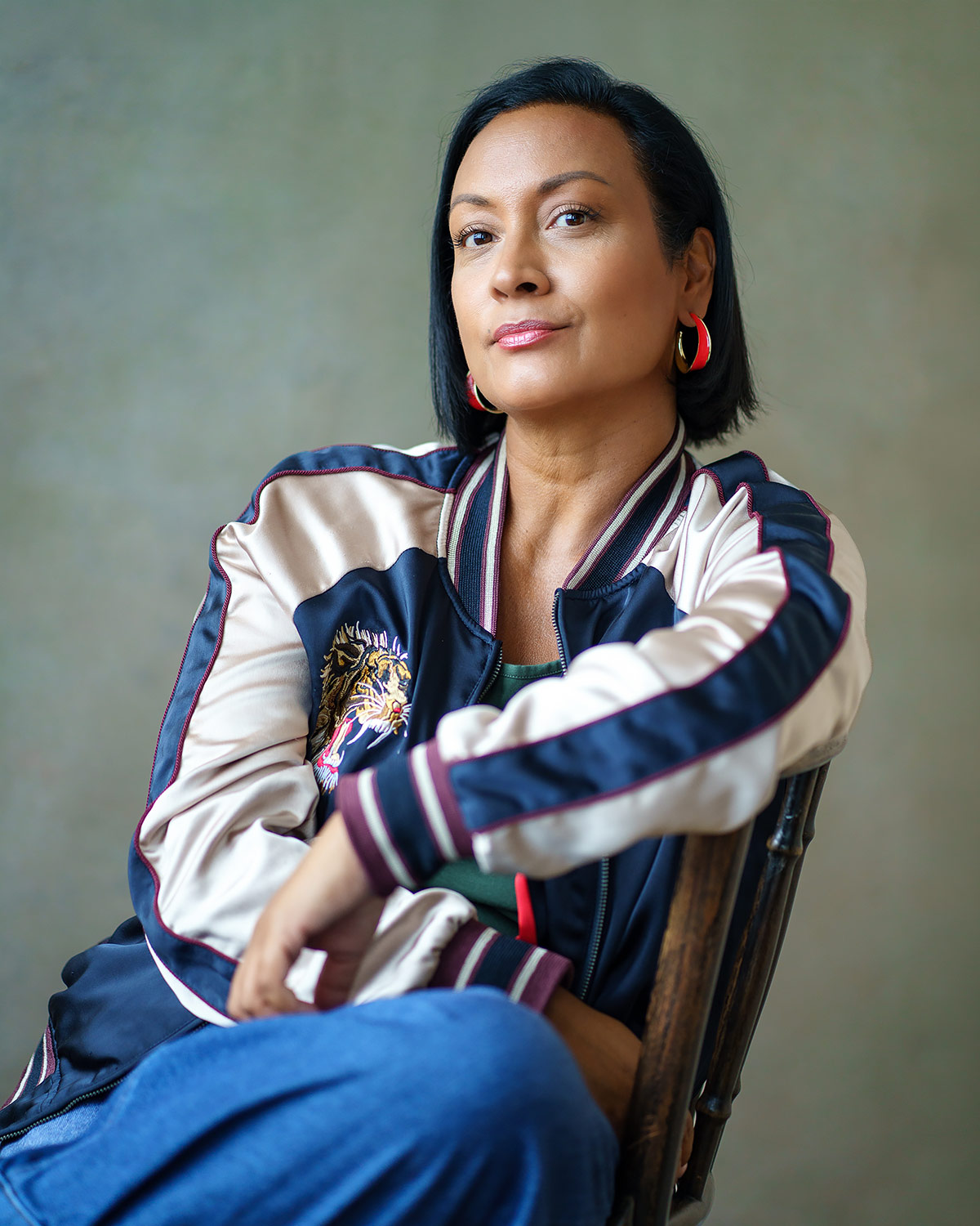 Vertical portrait of Theresa Godly seated on a wooden chair, wearing a navy and cream satin bomber jacket